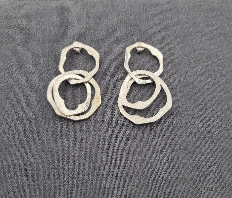 A pair of silver-toned geometric dangle earrings with overlapping textured rings, displayed on a dark gray textured fabric background.