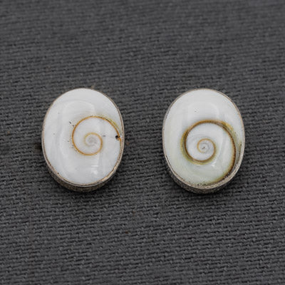 A pair of oval stud earrings featuring white mother-of-pearl or shell inlay with a spiral pattern in warm beige and brown tones, set in a silver-toned metal, displayed on a dark gray textured background.