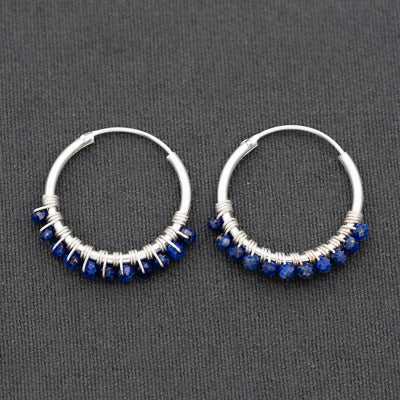 A pair of silver hoop earrings with a row of small faceted blue gemstones along the bottom curve, displayed on a dark gray textured fabric background.