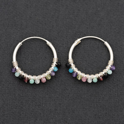 A pair of silver hoop earrings with small dangling gemstone beads in multiple colors, displayed on a dark textured background.