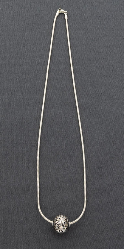 A silver-toned snake chain necklace with a single round, textured silver bead pendant, displayed against a dark gray background.