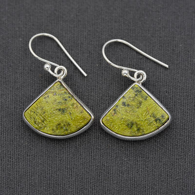 A pair of silver drop earrings with fan-shaped olive green stone pendants, displayed on a dark textured fabric background.