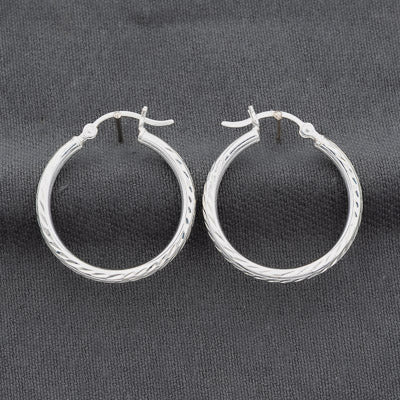 A pair of silver hoop earrings with a textured, twisted rope-like finish, displayed on a dark gray fabric background.